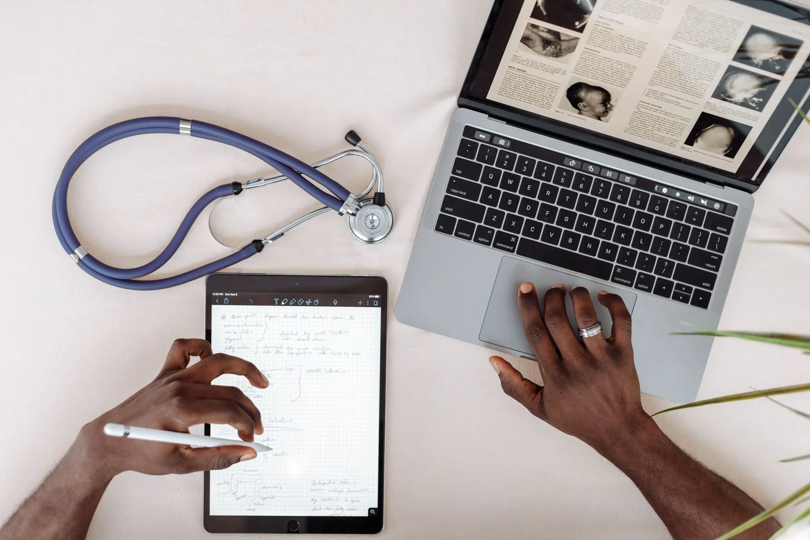 Overhead view of hands taking notes on a tablet beside a laptop showing a journal article and a stethoscope